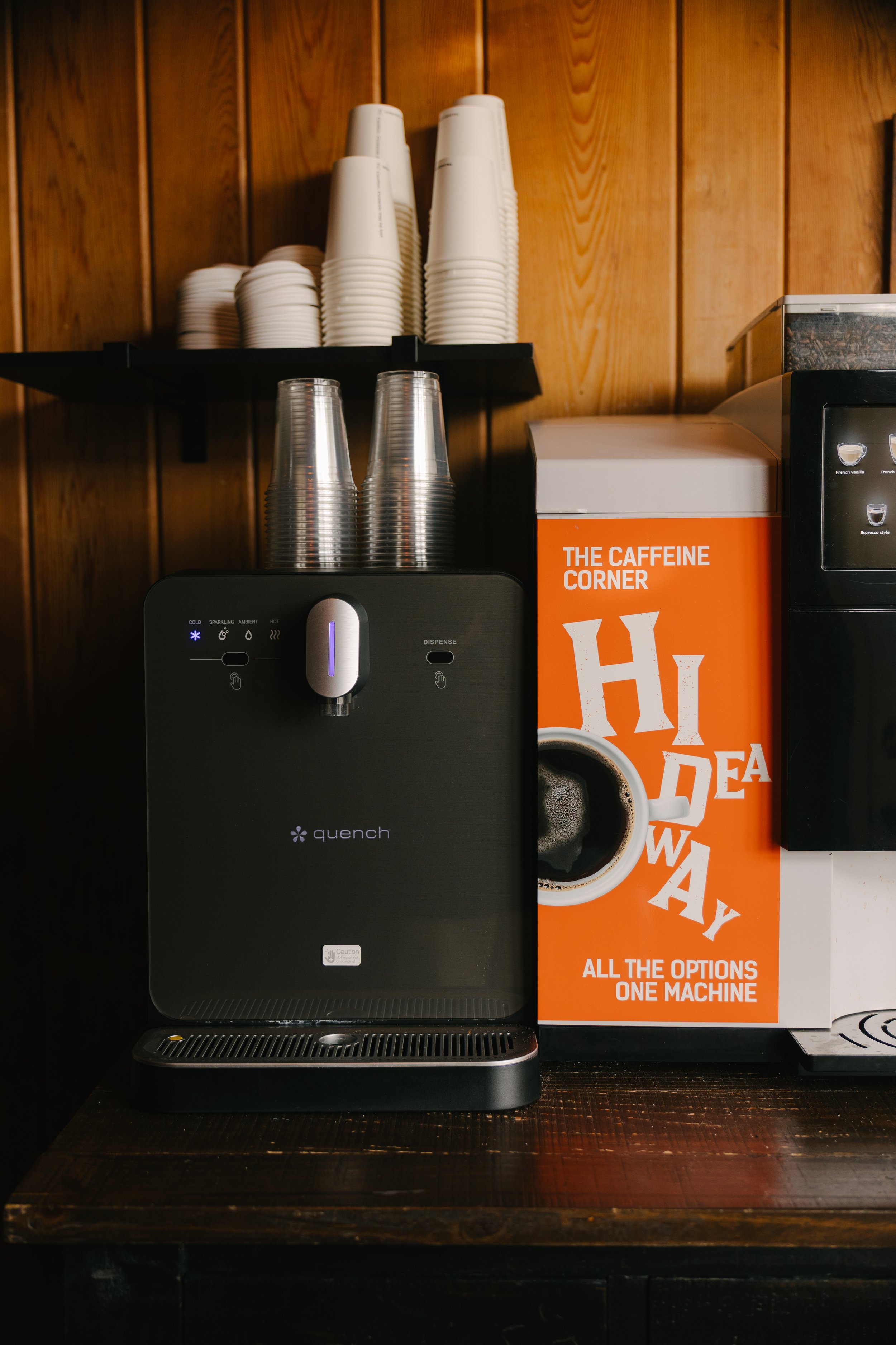 A coffee station with a Quench hot and cold beverage dispenser, disposable cups, and a box labeled 'The Caffeine Corner' on a wooden counter against a wood-paneled wall.