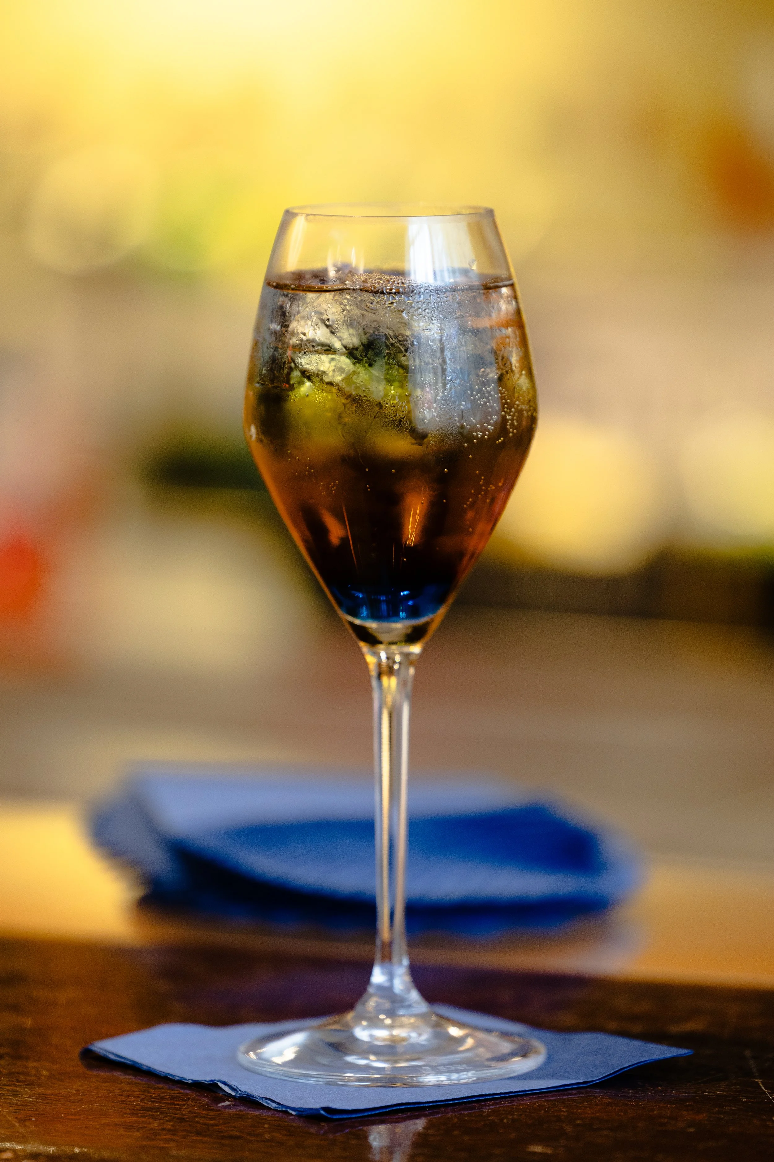 A glass of dark soda with ice cubes on a blue napkin on a wooden table.
