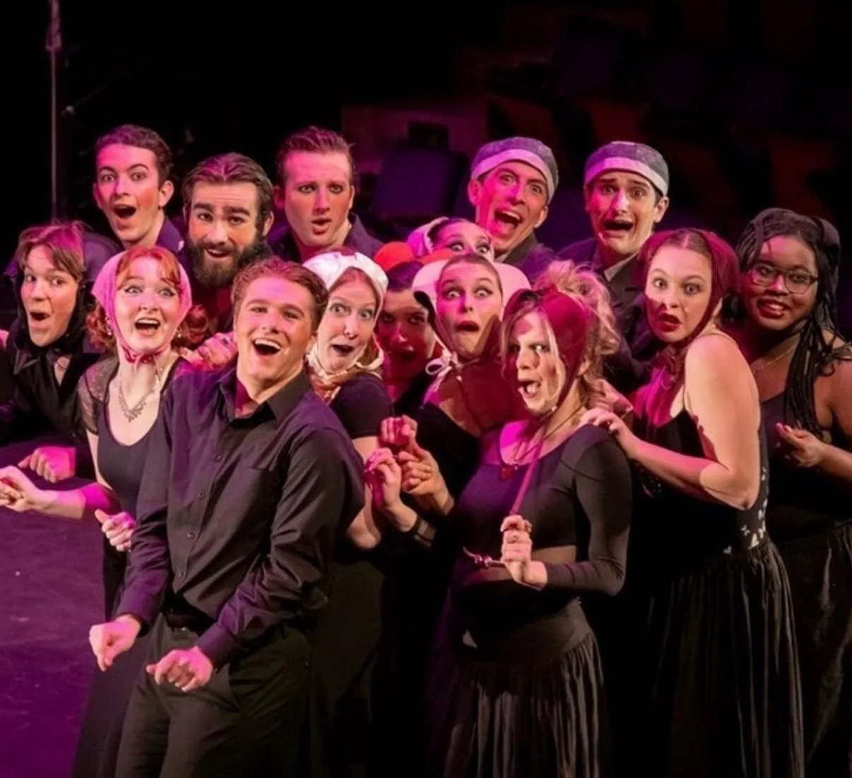 A group of actors on stage performing, all expressing surprise or excitement, with some wearing costumes and makeup, under theatrical lighting.