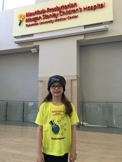 A girl wearing glasses, a black hat with a logo, and a yellow T-shirt standing inside a hospital. The background shows a sign reading 'Newport Presbyterian Morgan Stanley Children's Hospital Columbia University Medical Center.'