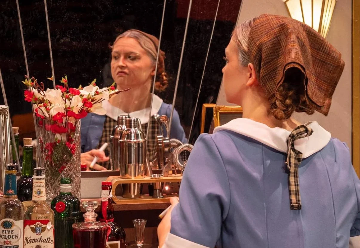 A woman dressed as a waitress with a retro outfit and headscarf looking into a mirror, with bottles and a vase of flowers on the bar counter.