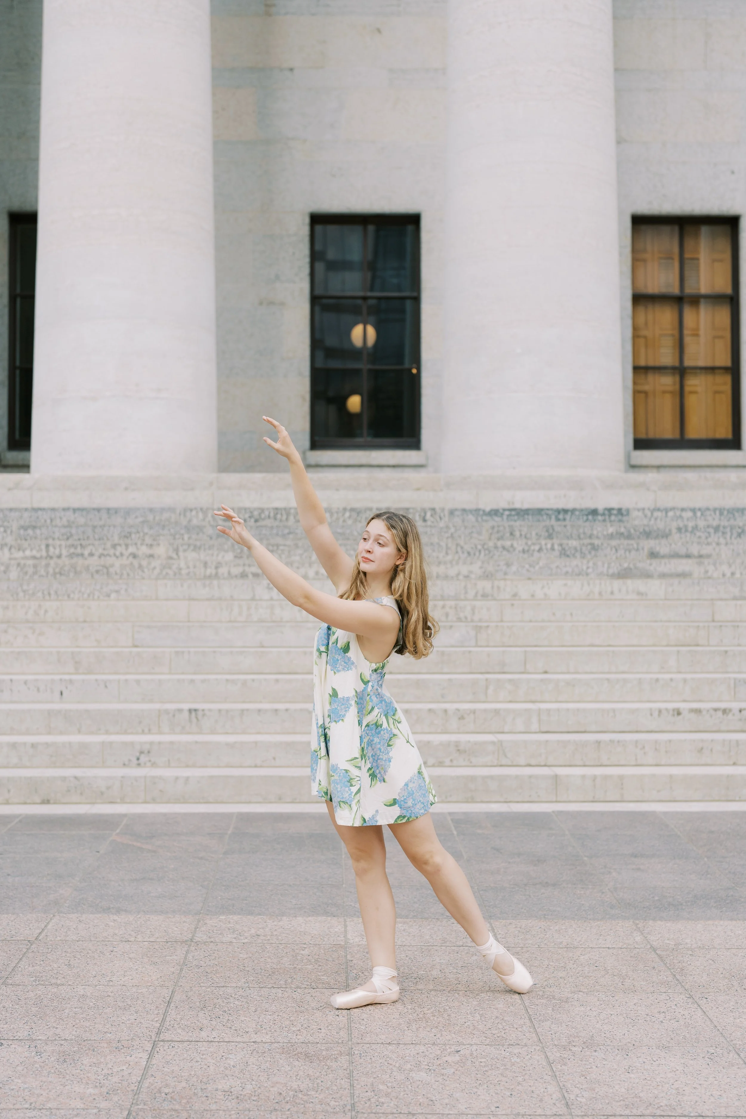 A young woman in a floral dance dress performing ballet outside in front of a large stone building with steps and tall columns.