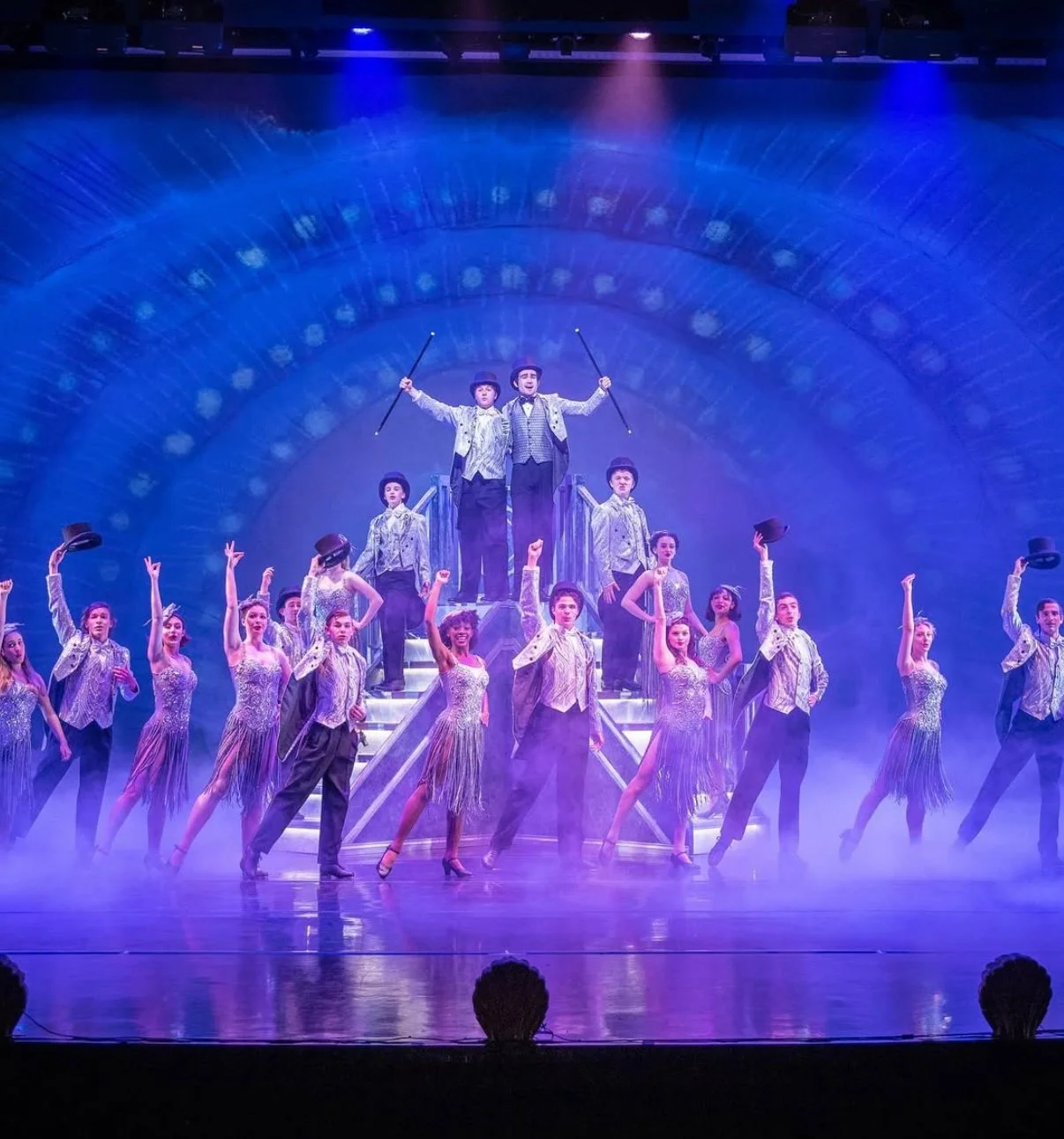 A theatrical stage performance with a large ensemble cast in vintage costumes, with two performers at the top holding canes, set against a vibrant blue and purple backdrop with fog.