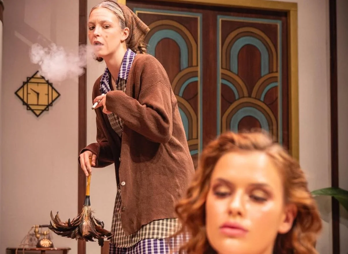 Woman smoking a cigarette and holding a broom in a room with brown, blue, and orange decor, including a colorful geometric door in the background, while another woman with curly hair is in the foreground.