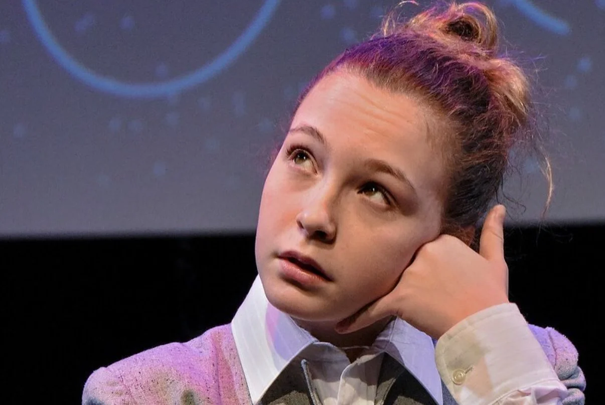 Young woman with reddish hair tied in a bun, resting her head on her hand, looking thoughtfully upward, with a dark background and faint blue circular designs.
