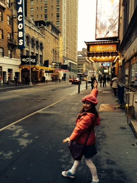 A young girl with a pink hat and coat, pointing down a busy city sidewalk lined with restaurants and hotels during sunset or dusk.