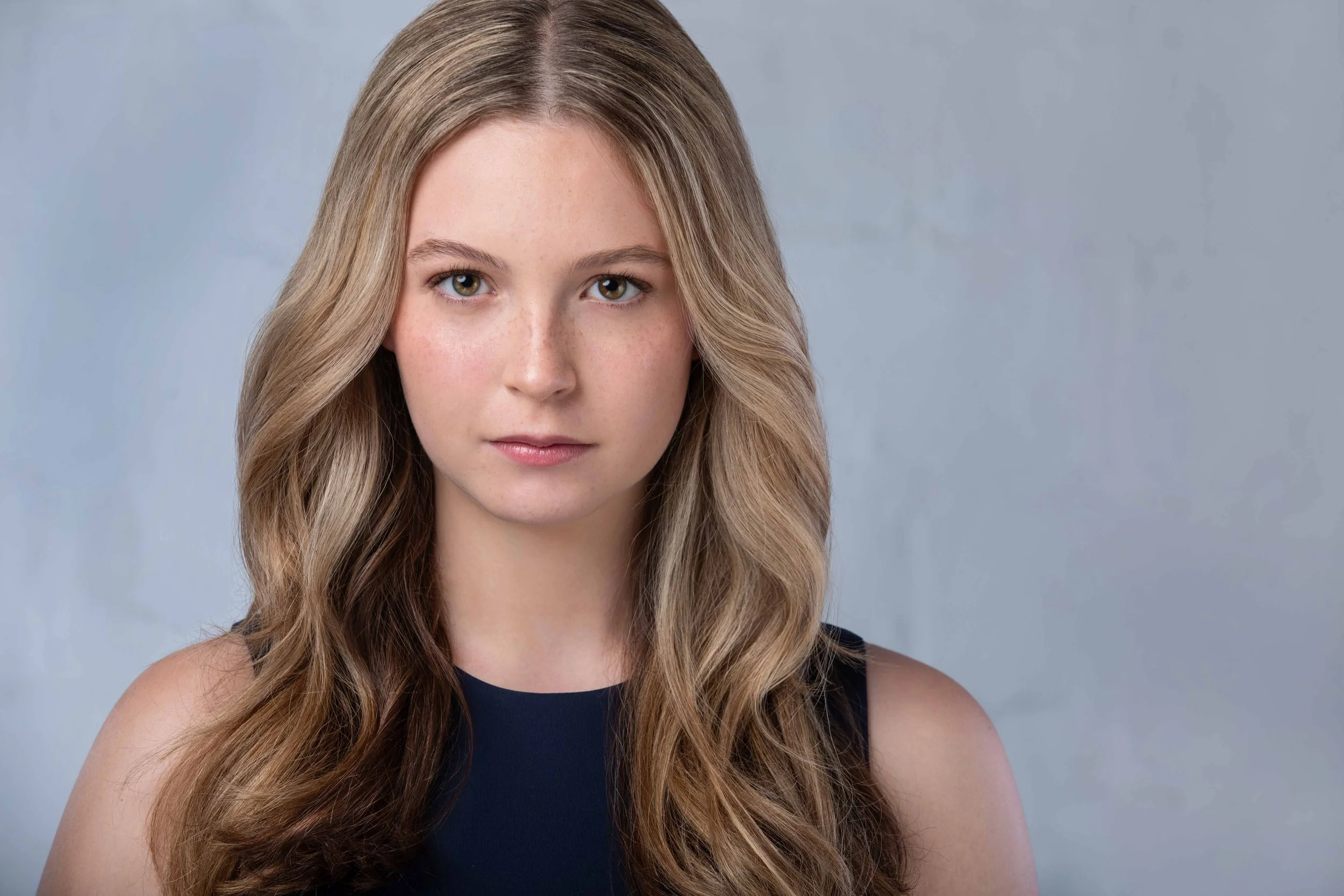 A young woman with long, wavy blonde hair, wearing a black sleeveless top, looking directly at the camera with a neutral expression, against a gray background.