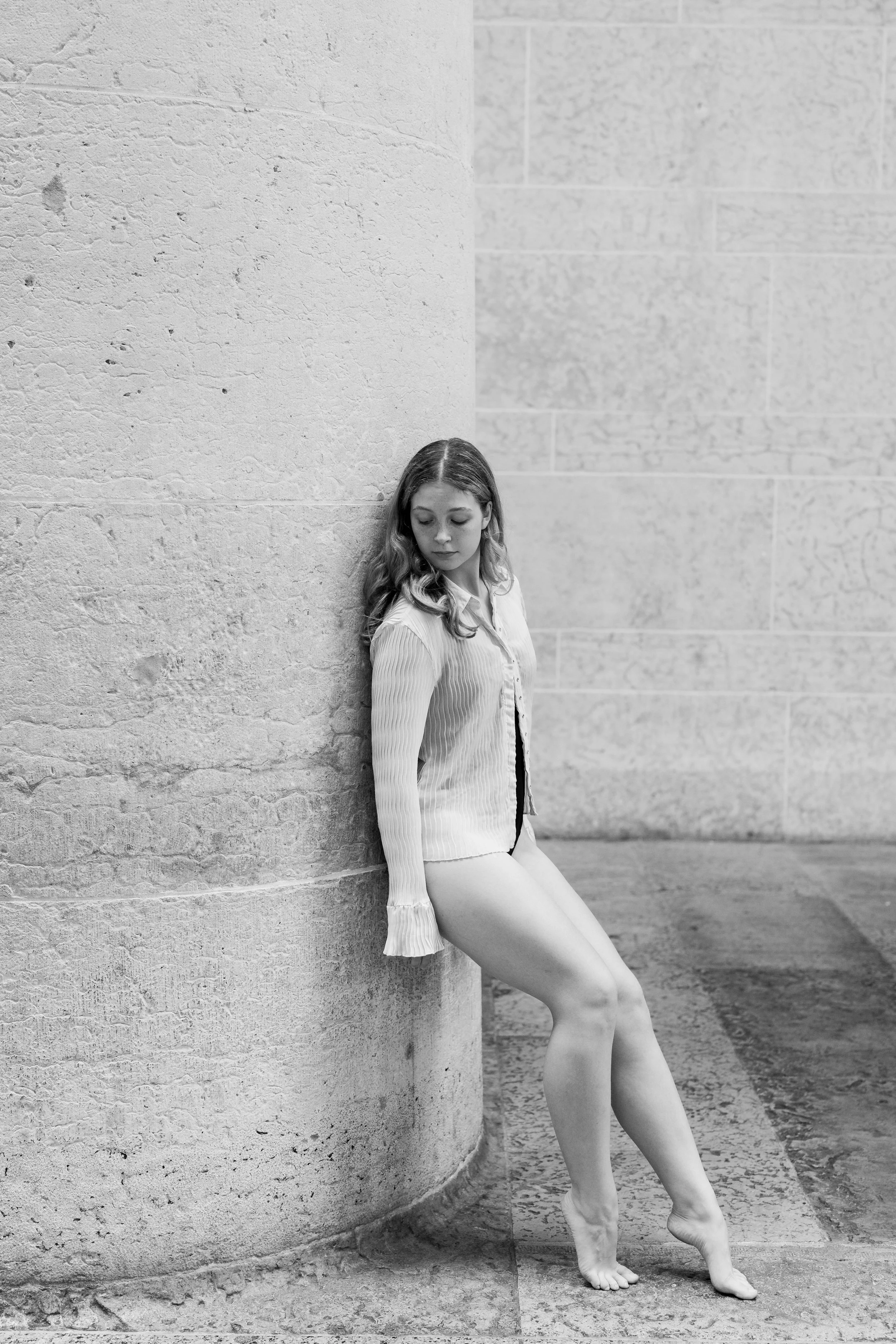 A black and white photo of a young woman with long wavy hair, sitting girl leaning against a large stone column, looking downward with a contemplative expression, indoors.