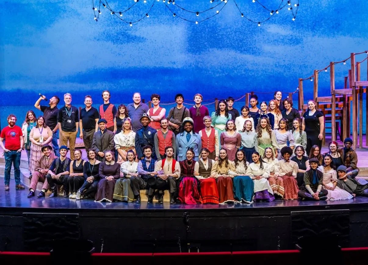 A large group of people on a stage, some sitting and some standing, with a painted backdrop of a blue sky and a wooden structure, under string lights, after a theatrical performance.