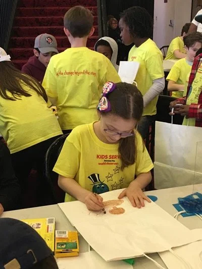 Children participating in a charitable event, with multiple kids in yellow t-shirts gathered around tables, some engaging in activities. Girl in front is writing or drawing on a large piece of paper.