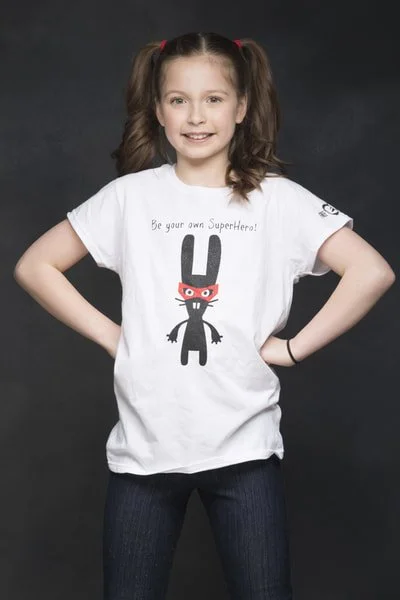Young girl with brown hair in pigtails, wearing a white T-shirt with a cartoon bunny and the text 'Be your own SuperHero!', standing with hands on hips against a dark background.