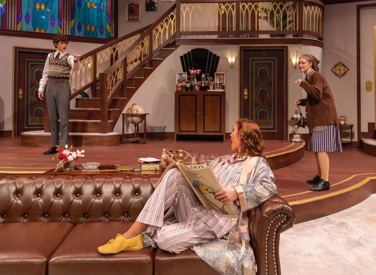 Scene from a play or TV show featuring a woman lounging on a leather couch reading a newspaper, two women standing and talking on a wooden stage with a staircase and vintage decor in the background.