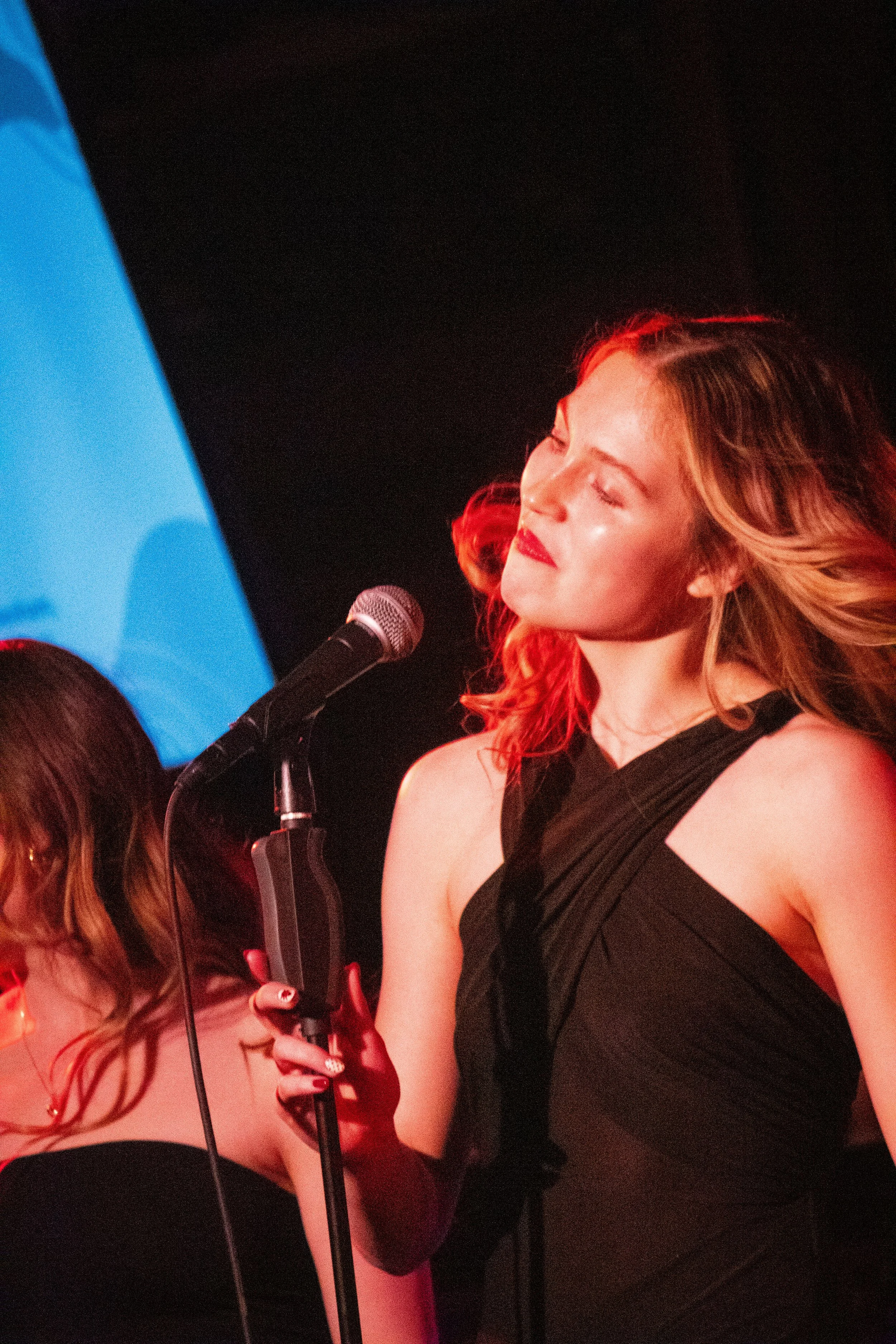 A woman singing into a vintage-style microphone with a black dress, closed eyes, and wavy hair, on stage under red lighting.