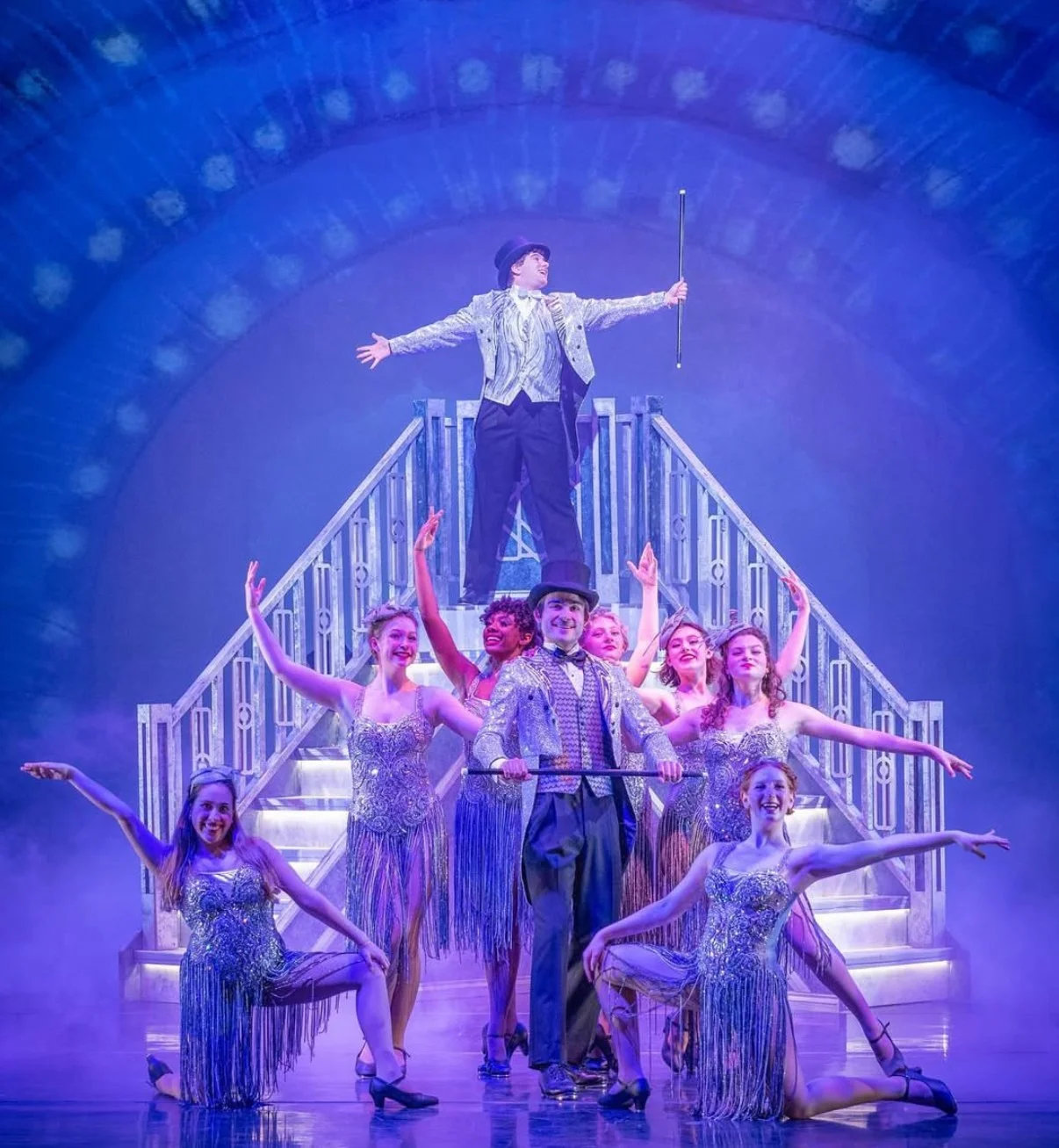 Theatrical stage scene with a male performer standing half-naked on a staircase with arms outstretched, surrounded by female dancers in glittery costumes, with a male performer in a top hat and coat holding a cane in front, under blue lighting.