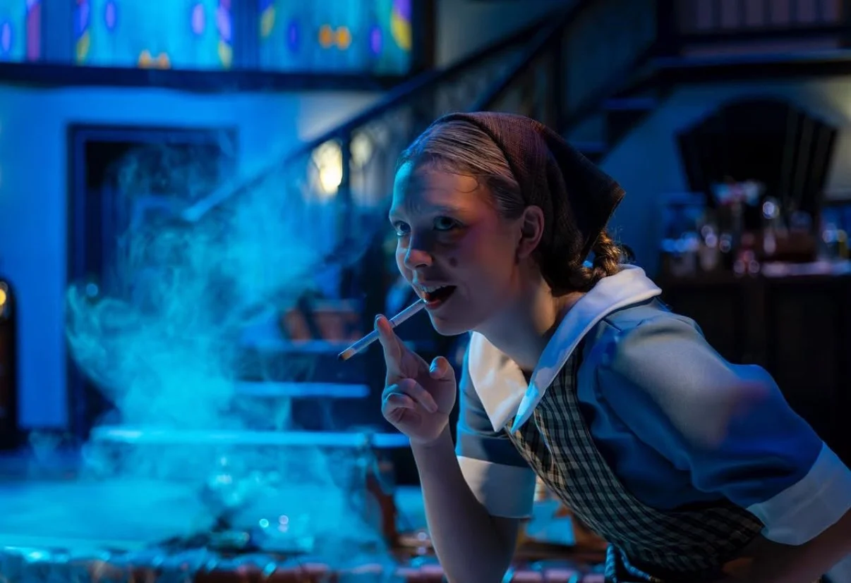 Woman in a checkered apron and headscarf blowing smoke from a cigarette in a dimly lit room with a blue hue.