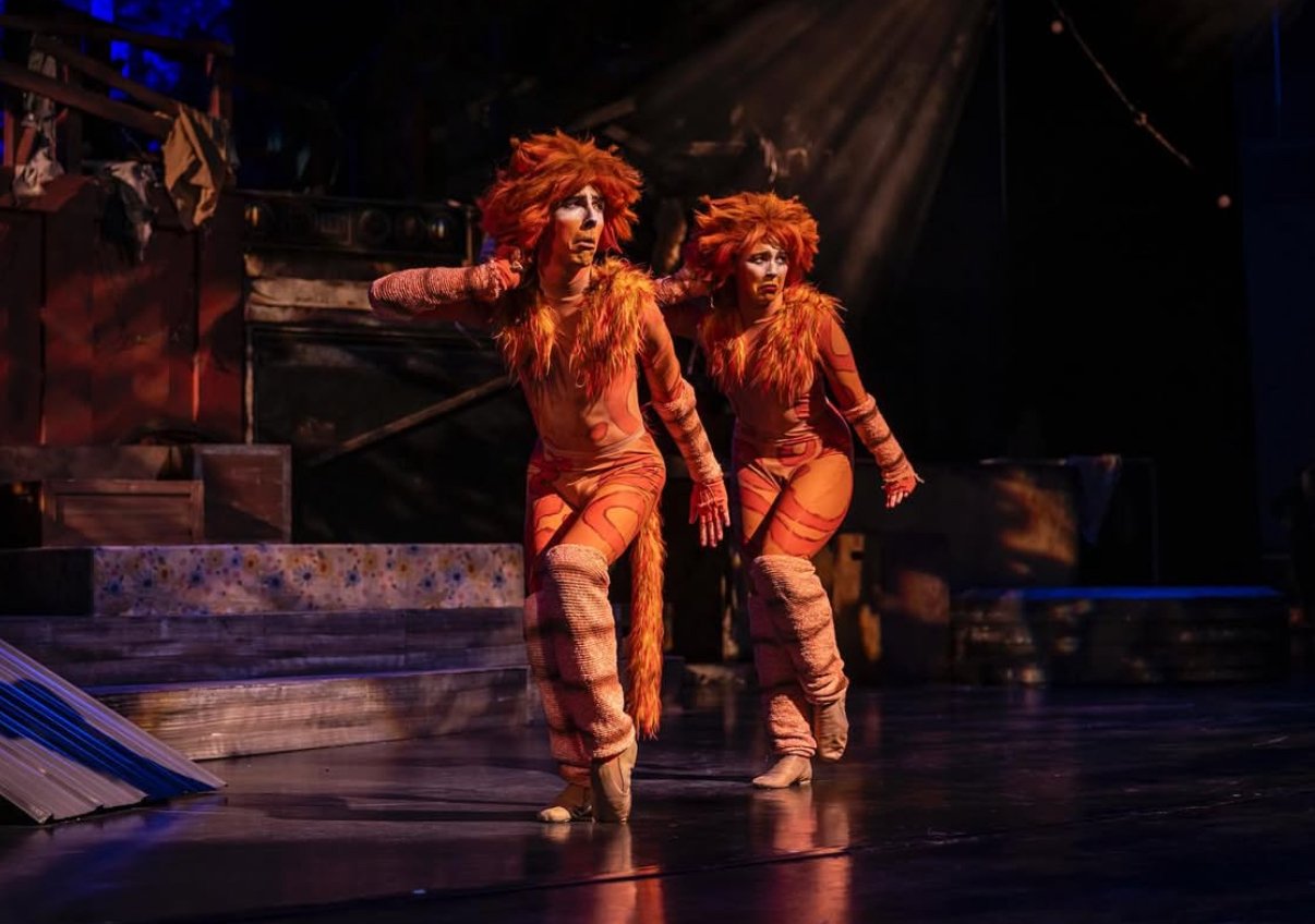 Two performers dressed as lions with orange fur and matching costumes on a stage during a theatrical performance.