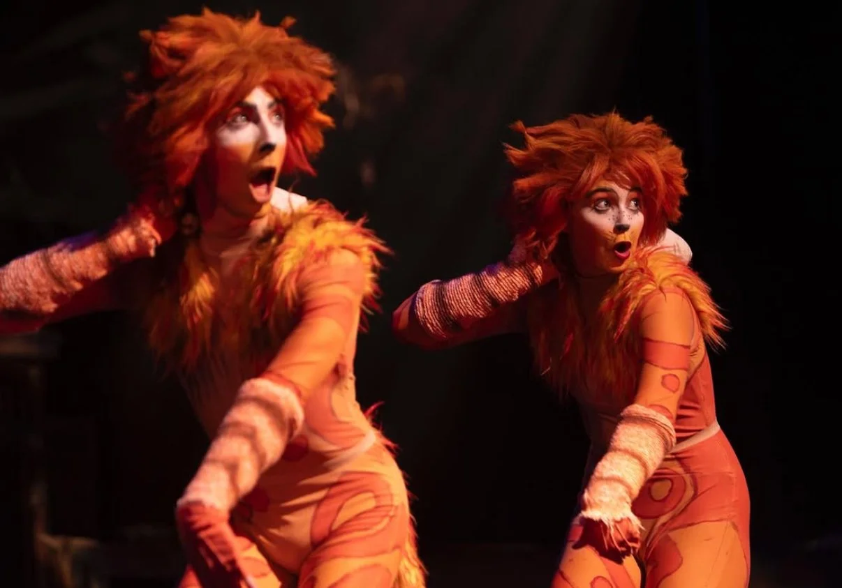 Two performers dressed as lions with orange and brown fur costumes and face makeup, appearing surprised or reacting to something on stage in a theatrical performance.