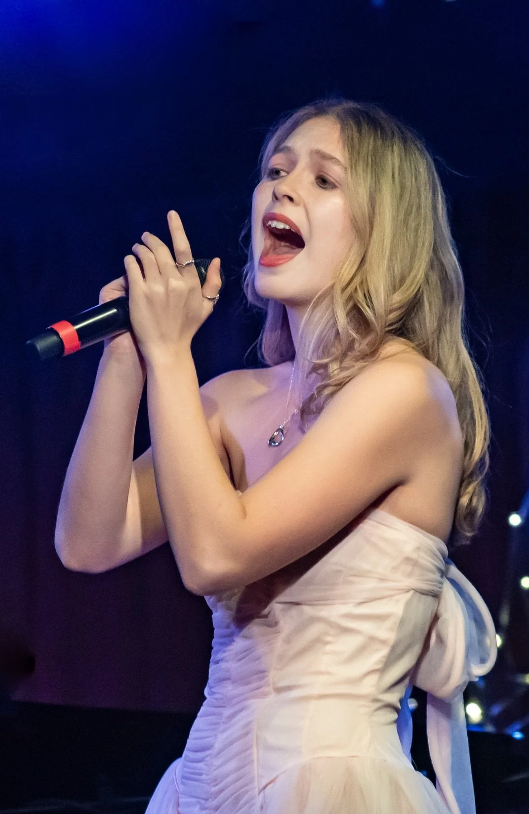 A young woman singing into a microphone during a performance, wearing a strapless dress and a necklace, with a dark background.