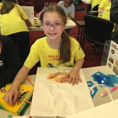Girl with glasses sitting at a table with drawing materials, smiling, in a classroom or activity setting.