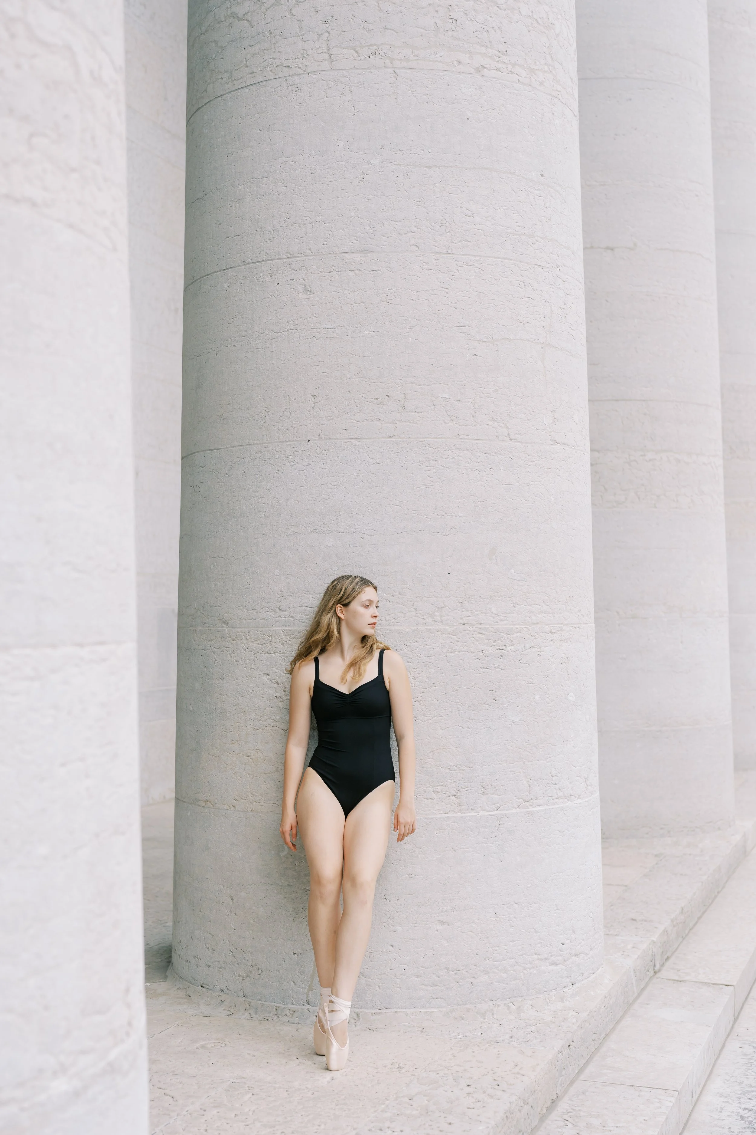 A ballerina in a black leotard standing on pointe shoes next to large stone columns.