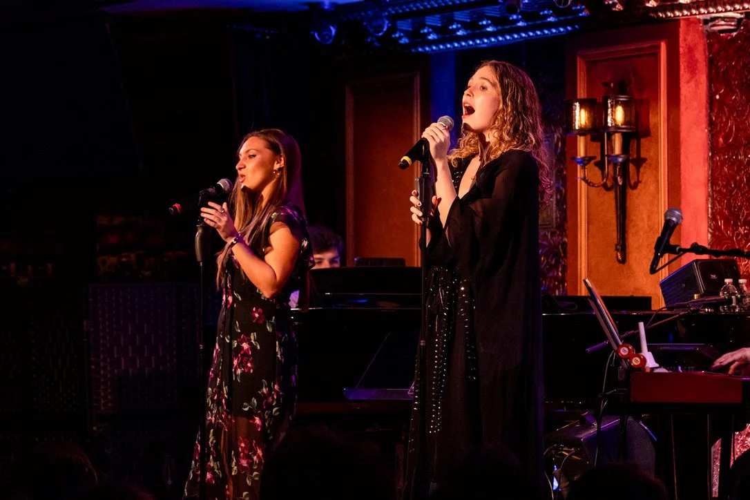 Two women singing with microphones on a stage, with a pianist in the background, in a cozy, warmly-lit performance venue.