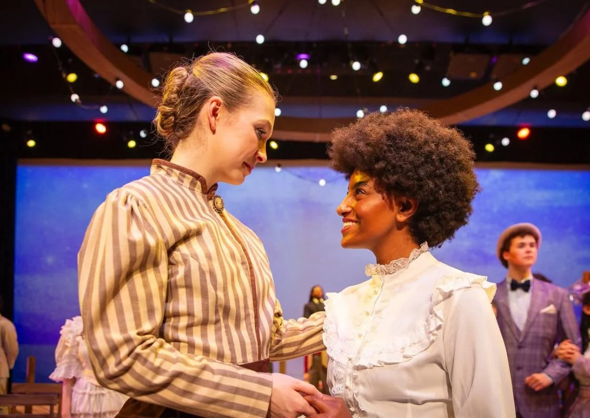 Two women in vintage clothing holding hands and smiling at each other on stage, with other actors in period costumes in the background. The stage is decorated with string lights and a blue backdrop.
