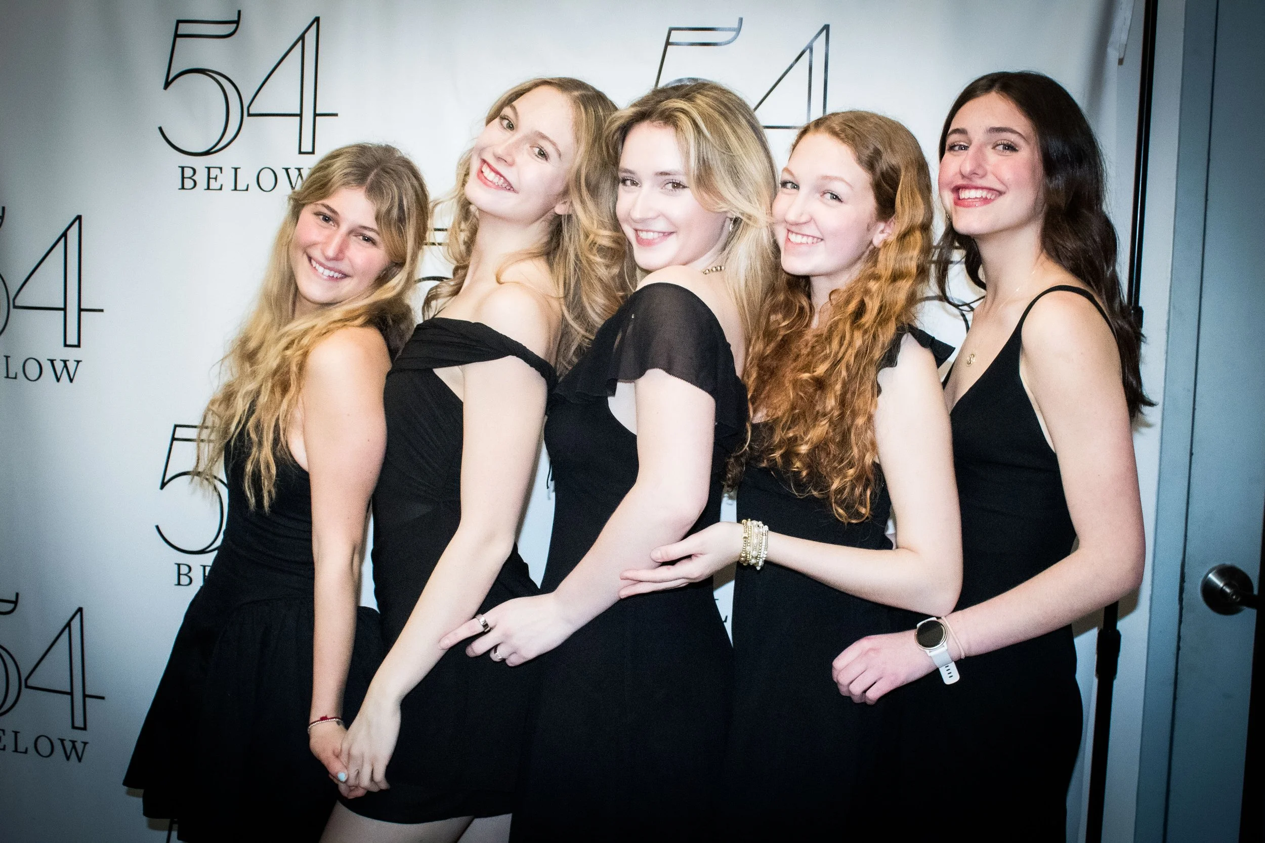 Five young women standing close together, all dressed in black dresses, smiling at the camera against a backdrop with numbers and the word 'below'.