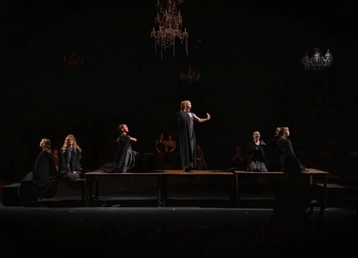 The image shows a theatrical performance with six women dressed in black, positioned on and around a rectangular stage. The woman in the center is standing, gesturing with her hand, while the others are sitting or kneeling on the stage. The backgroun