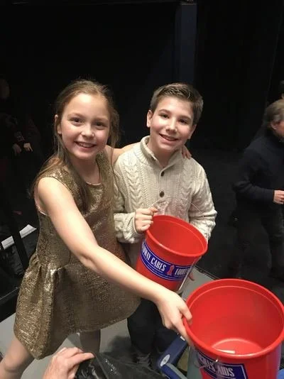 Two children, a girl and a boy, smiling and holding red buckets at an indoor event.
