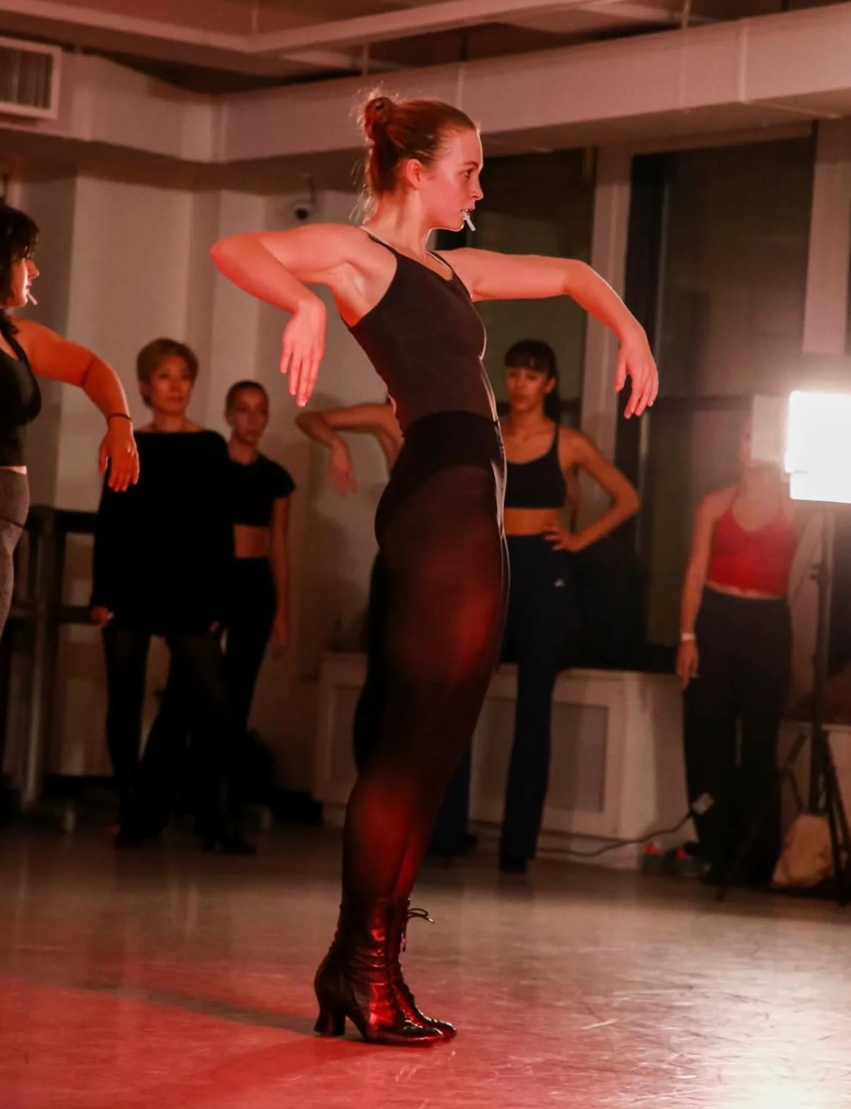 A woman in black dance attire and high-heeled boots performs a dance routine in a dimly lit studio with several women watching in the background.
