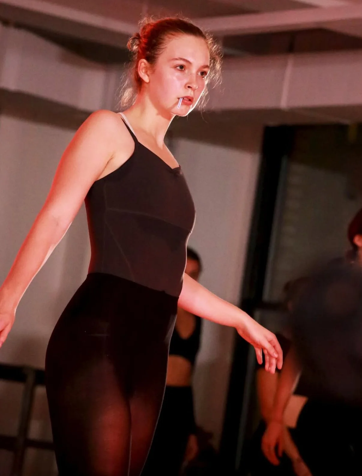 A young woman with curly hair tied back, wearing a black sleeveless top and sheer black tights, standing with her arms outstretched and a toothpick in her mouth, in an indoor setting.