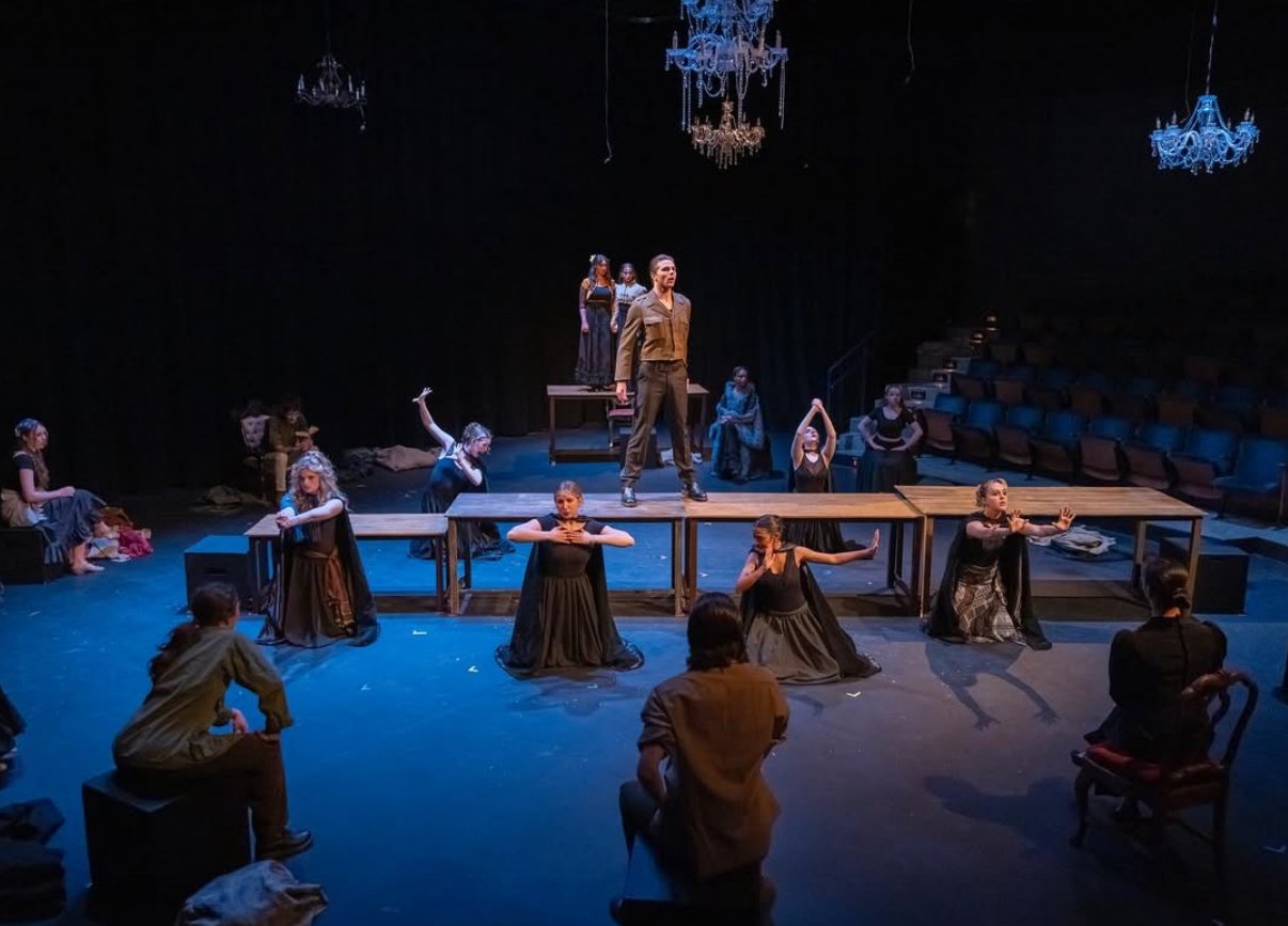 The scene shows a theatrical performance with actors on stage, some standing on elevated platforms and others kneeling or sitting on the stage floor. The stage is decorated with chandeliers hanging from the ceiling, and the background is dark, emphas