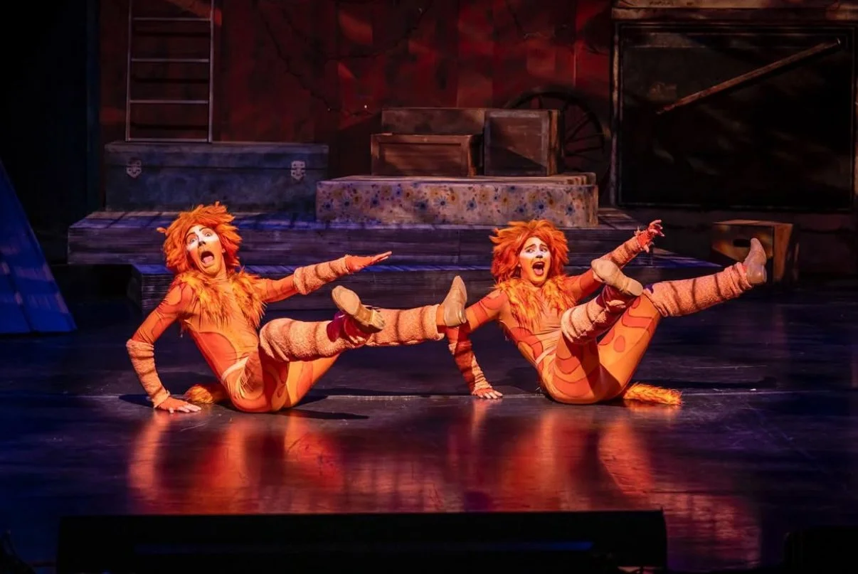 Two performers in lion costumes performing on stage with their legs raised and mouths open, in a theatrical production.