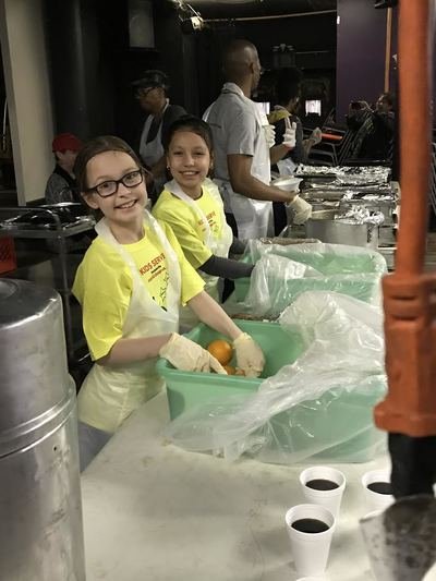 Two young girls smiling with gloves on, volunteering at a food distribution event, surrounded by volunteers and supplies.
