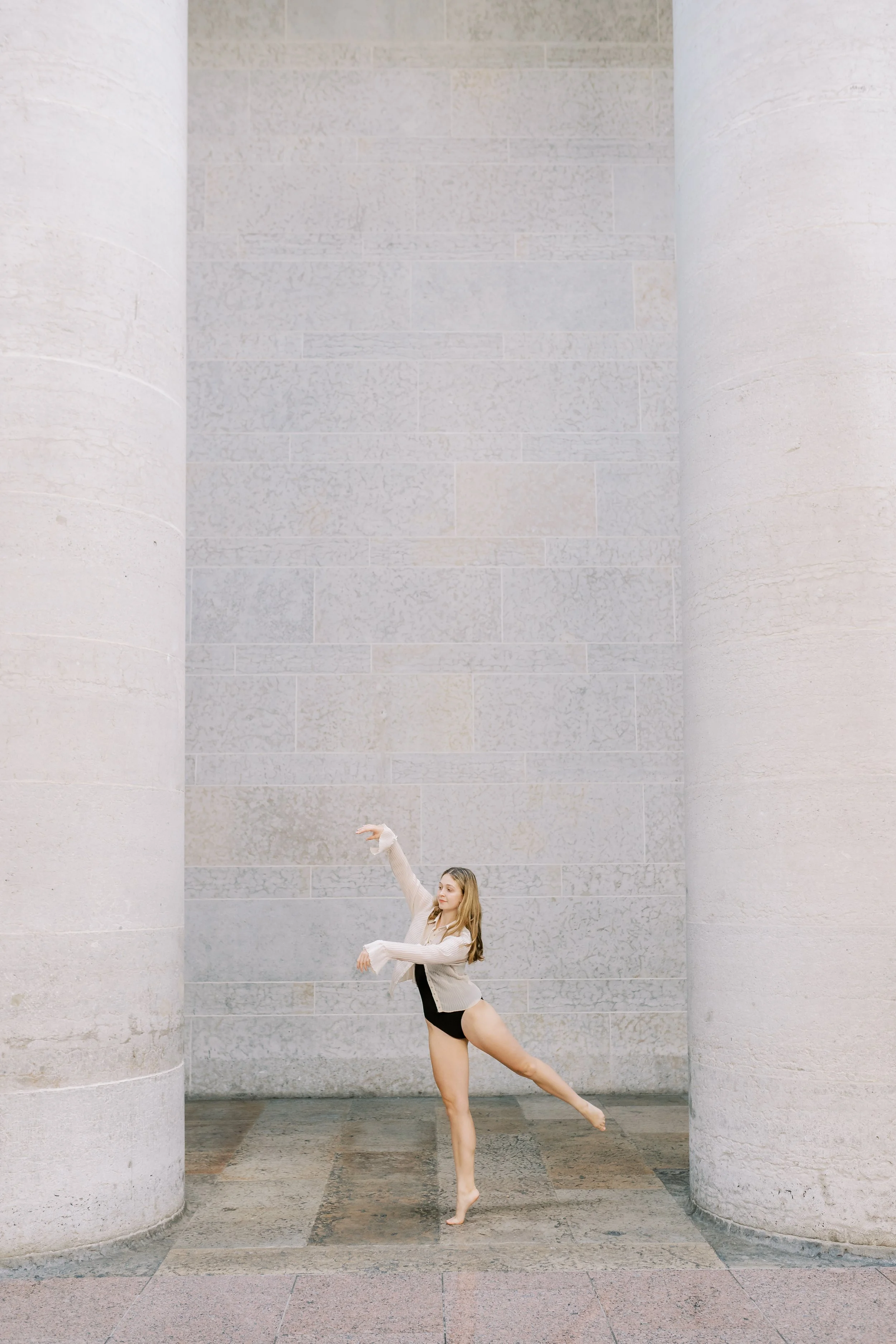 A young woman practicing ballet or dance in a spacious corridor with tall stone columns and a light-colored brick wall.