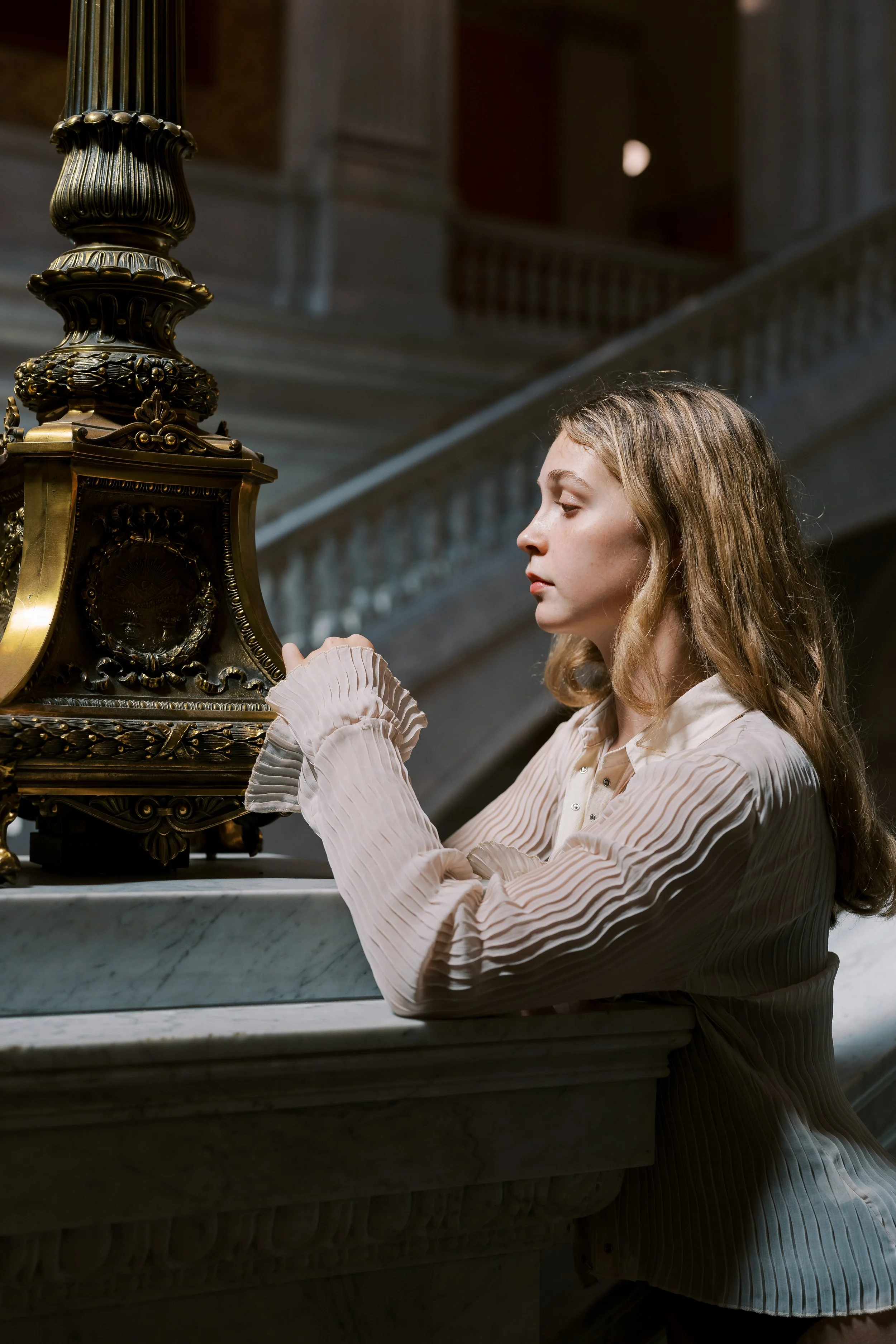 A young woman with wavy blonde hair sitting at a marble counter, looking at an ornate, antique brass clock in a grand, historic building.