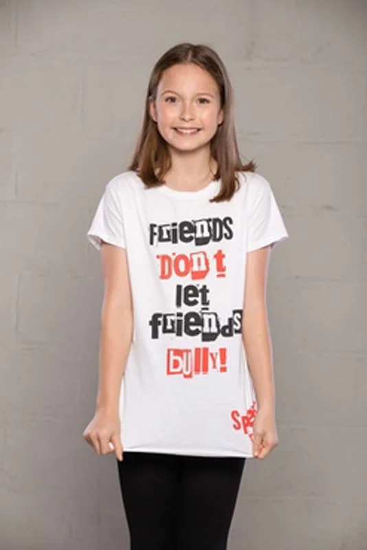 A young girl with shoulder-length brown hair smiling, wearing a white T-shirt that says 'Friends don't let friends bully!' in black and red letters and black pants, standing against a plain gray background.