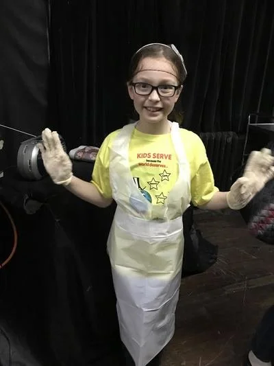 Young girl wearing glasses, a yellow T-shirt, a plastic apron, and gloves, participating in a community service event with a black curtain background.