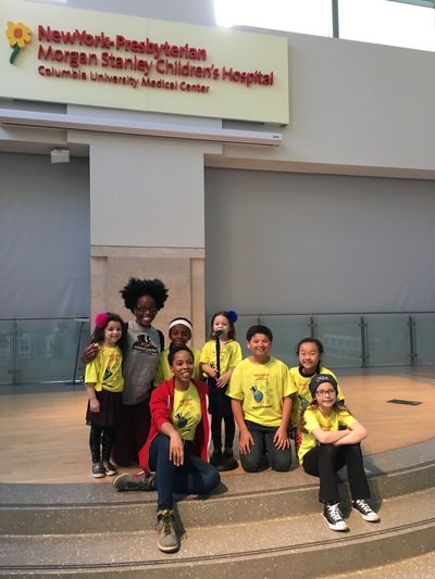 A group of six children and one adult pose in front of a hospital sign at Columbia University Medical Center. The children wear yellow shirts, and the adult is dressed in casual clothes.