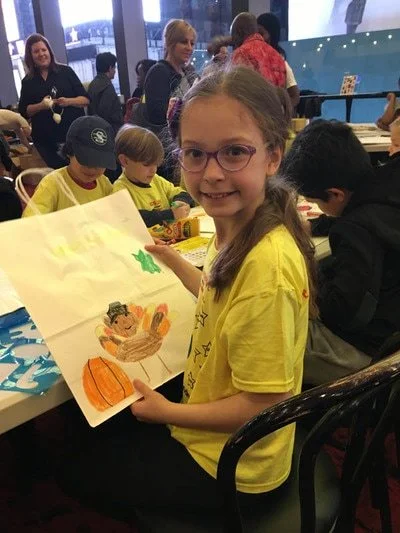 A young girl with glasses and a yellow shirt, smiling and holding a completed craft or drawing featuring a turkey, a pumpkin, and a Christmas tree, at a craft event with other children and adults in the background.