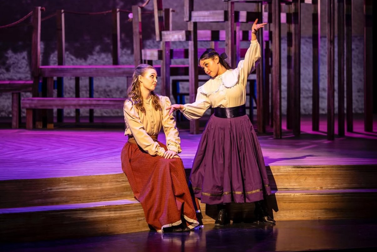 Two women singing on a theater stage, one sitting and the other standing, surrounded by a wooden set with pink and purple lighting.