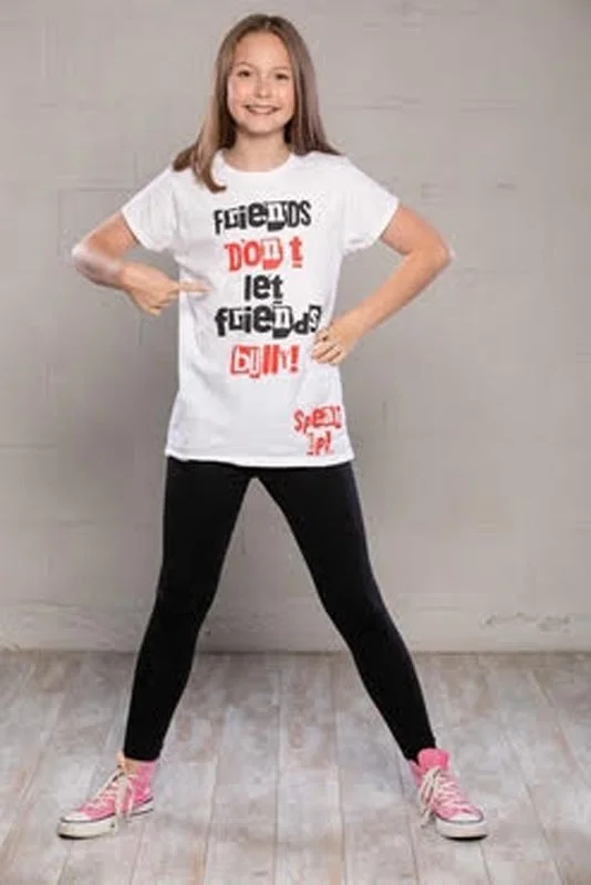 Young girl pointing at her white T-shirt with red and black text that says 'Friends don't let friends go in!'. She is smiling, wearing black leggings and pink sneakers, standing on a wooden floor against a plain gray wall.
