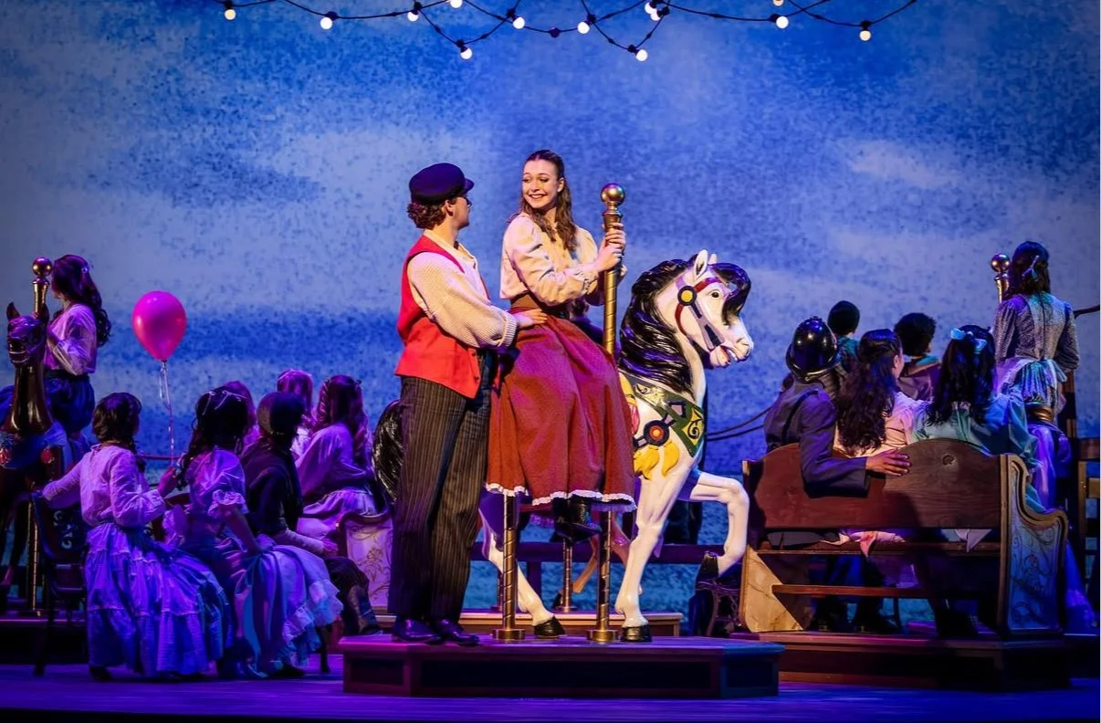 Children performing in a scene from a musical or school play, with a young girl on a carousel horse holding a staff, surrounded by children in costume and audience members.
