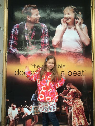 Young girl standing in front of a movie poster with two adults. The girl is smiling and giving two thumbs up, wearing a pink and white patterned jacket and a floral skirt.