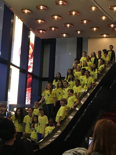 Group of children and adults wearing yellow shirts, ascending an escalator inside a modern building with a high ceiling and large windows.