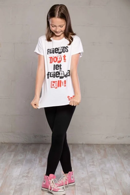 A smiling girl wearing a white T-shirt with a printed message, black leggings, and pink sneakers, standing against a plain wall.