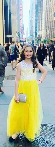 A young girl in a yellow and white dress standing on a busy city street with tall buildings and crowds of people.