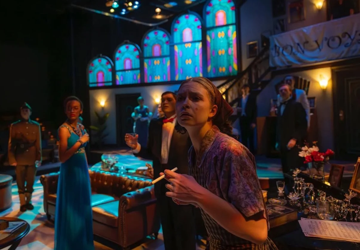 A woman with short hair holding a cigarette, looking distressed in a dimly lit room with colorful stained glass windows and several people in formal attire in the background, possibly at a party or performance.