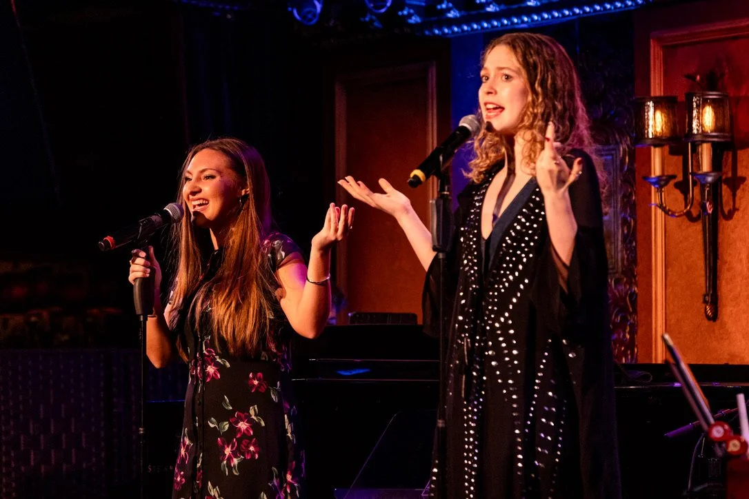 Two women singing into microphones on a stage with a dark and ornate background.