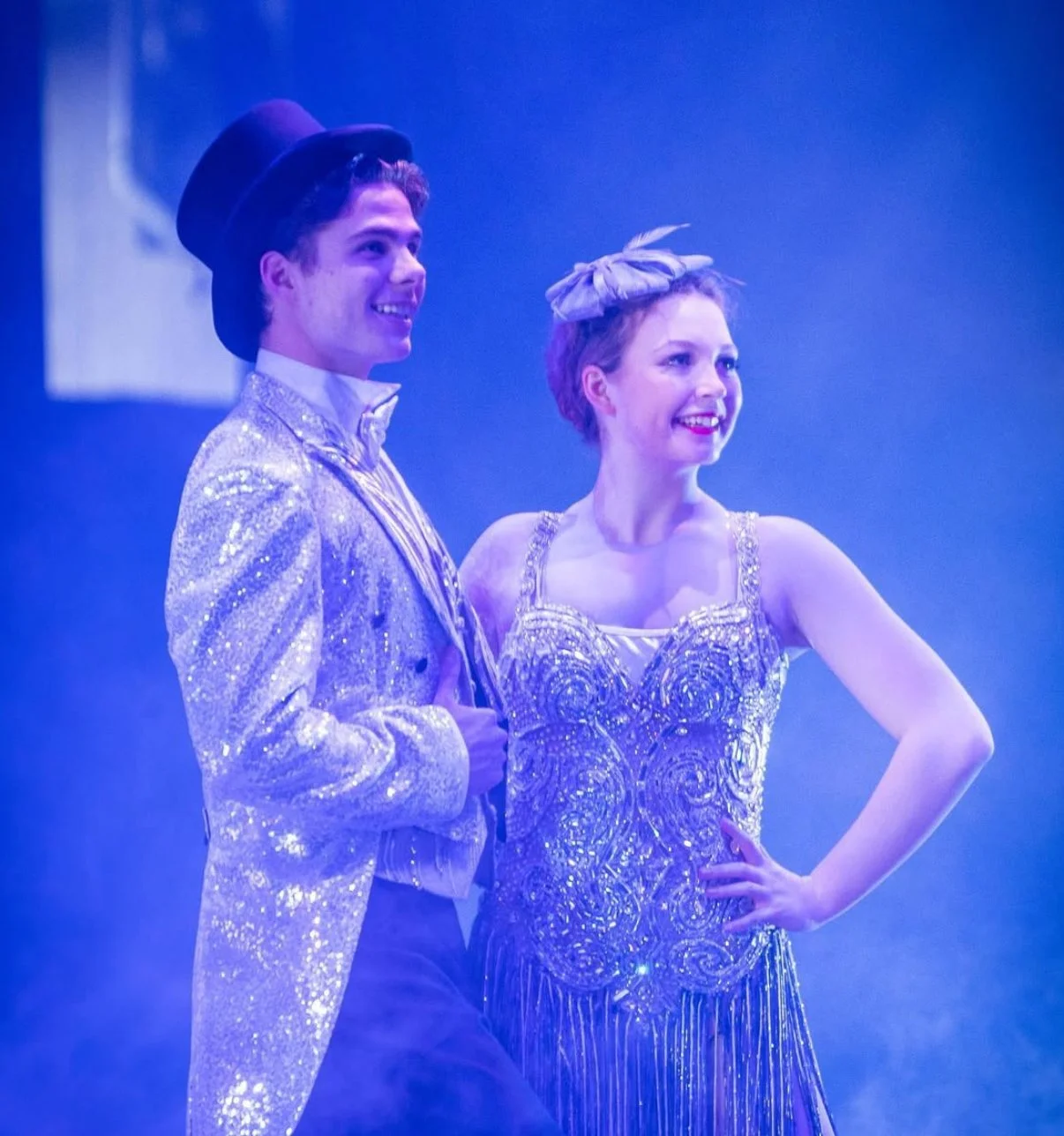 Two performers in shiny costumes standing together, one male and one female, smiling under purple stage lighting.